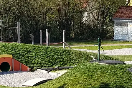 Ein Kinderspielplatz mit Tunnel, Rutsche, Schaukel und Wasserpumpe. Der Spielplatz ist von grünem Rasen und Bäumen umgeben.