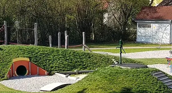 Ein Kinderspielplatz mit Tunnel, Rutsche, Schaukel und Wasserpumpe. Der Spielplatz ist von grünem Rasen und Bäumen umgeben.