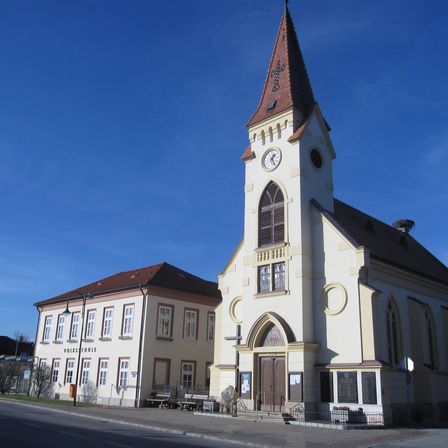 Eine große Kirche mit einem braunen Turm und einer Uhr, neben einem Schulgebäude mit vielen Fenstern unter einem klaren blauen Himmel.