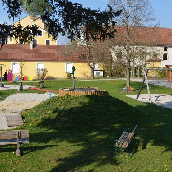 Ein Spielplatz mit Sandkasten, Wasserpumpe, Trampolin und Schaukel befindet sich vor einem gelben Gebäude mit farbenfrohen Zeichnungen.