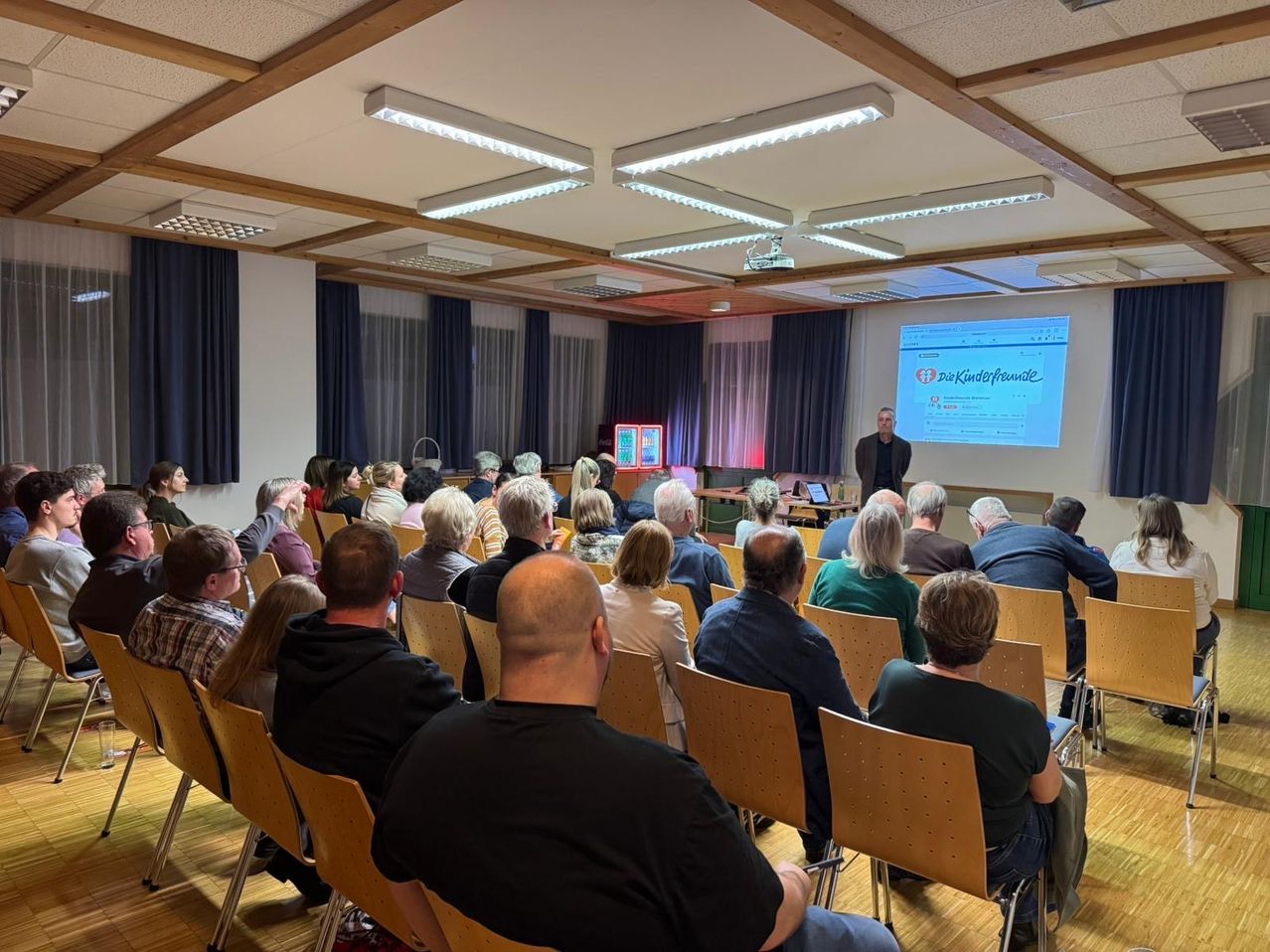 Ein Mann steht vor einer Projektionsleinwand und präsentiert einem sitzenden Publikum in einem Konferenzsaal. Der Raum hat Holzböden und -decken, und mehrere Lampen beleuchten den Raum.