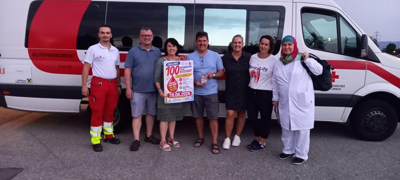 Eine Gruppe von Menschen steht vor einem Bus und posiert lächelnd für ein Foto. Eine Frau hält ein Schild mit dem Text 'Challenge 100' und einem Bluttropfen-Symbol. Das Datum '28.08.2026' steht ebenfalls auf dem Schild.