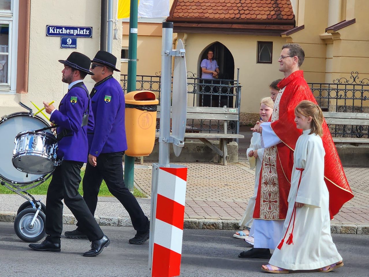 Zwei Männer in Lila und Schwarz gehen an einem Mülleimer vorbei. Ein Priester in roten und weißen Gewändern geht mit zwei Kindern. Eine Frau steht in der Tür einer Kirche.