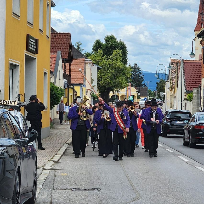 Eine Gruppe von Musikern in lila Outfits und Hüten spielt Instrumente bei einem Straßenumzug. Ein Mann in einem schwarzen Anzug steht am Straßenrand. Autos sind auf beiden Seiten der Straße geparkt.