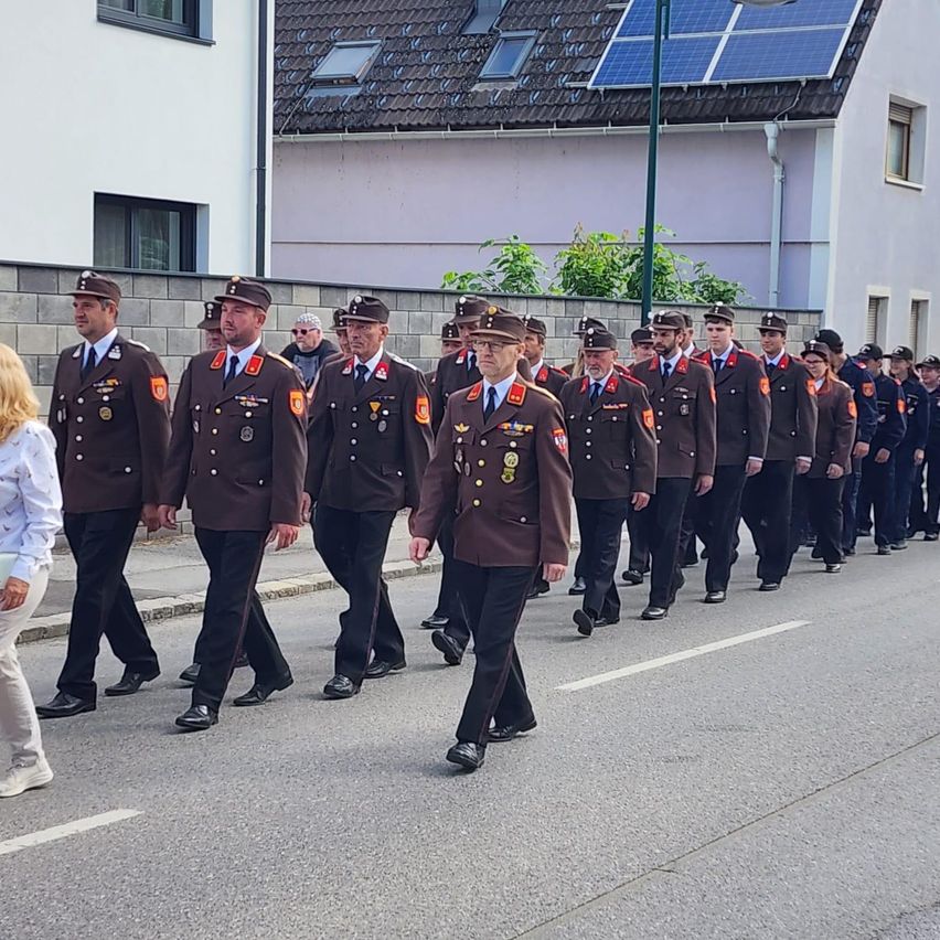Eine Gruppe uniformierter Personen marschiert die Straße entlang, begleitet von Zuschauern und Gebäuden mit Solarpanelen.