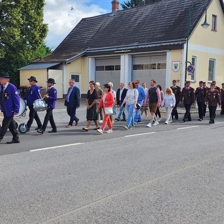 Eine Gruppe von Menschen in Uniformen geht auf einer Straße, begleitet von Trommlern. Sie sind vor einem Gebäude mit Schornstein und zwei Garagen.