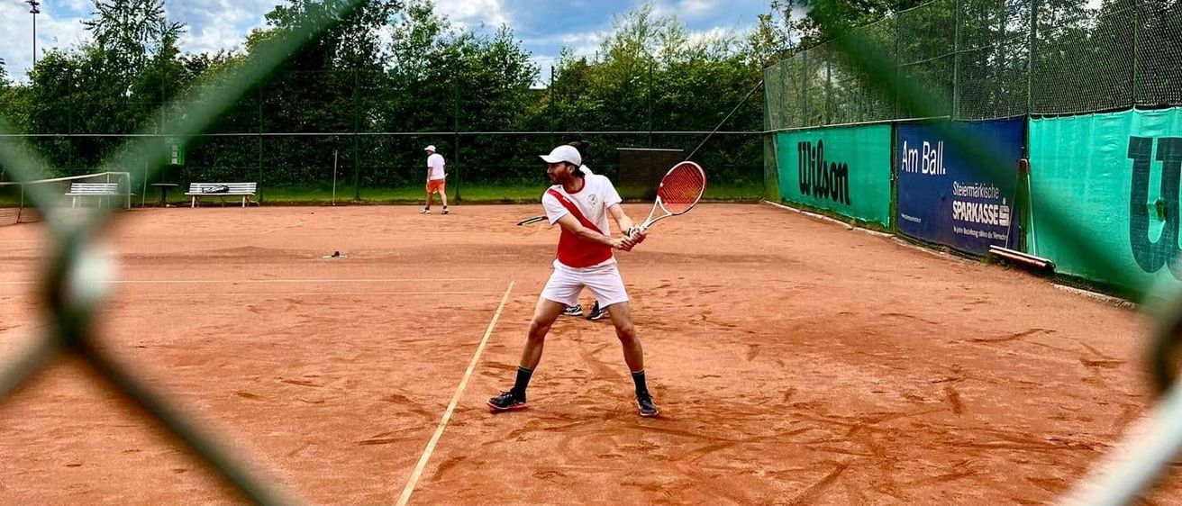 Ein Tennisspieler schwingt seinen Schläger auf einem Sandplatz. Im Hintergrund sind ein weiterer Spieler und ein Netz zu sehen. Die Szene wird von einem Zaun mit Banner eingerahmt.
