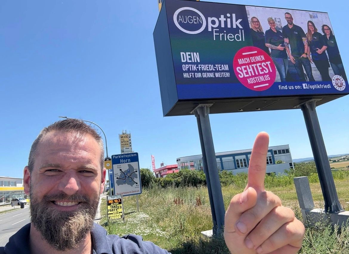 Ein Mann gibt vor einem großen Werbeschild mit dem Team von Augen Optik Friedl und einem kostenlosen Sehtestangebot einen Daumen hoch. Dahinter befindet sich ein Grasfeld mit Straßenschildern.