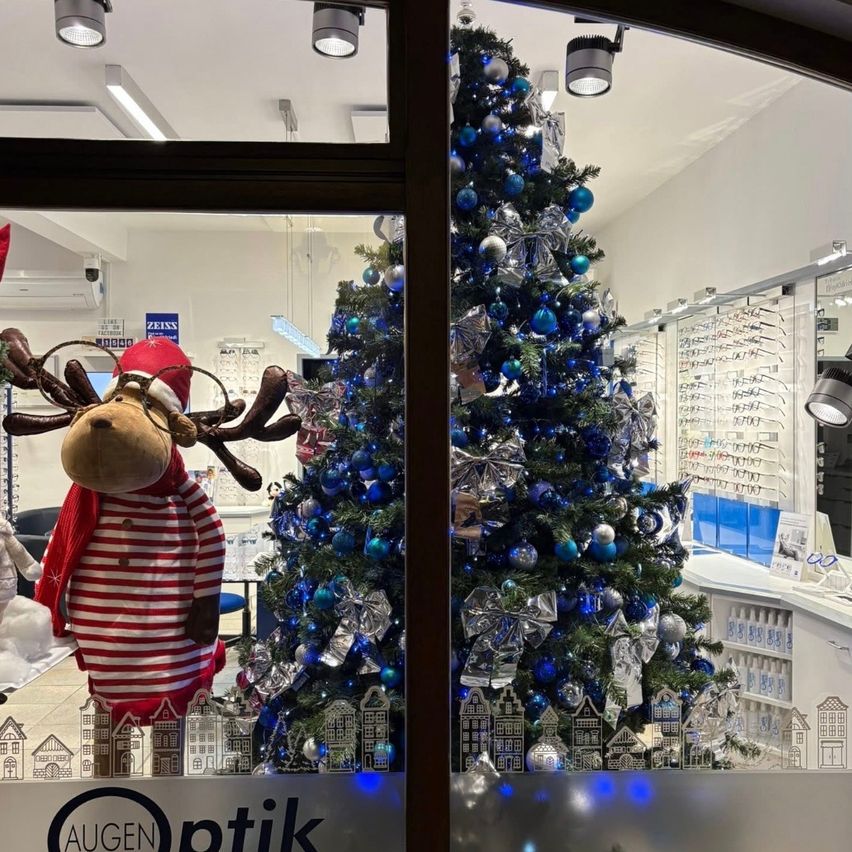 Ein mit blauen und silbernen Kugeln geschmückter Weihnachtsbaum ist im Schaufenster eines Geschäfts zu sehen. Ein weihnachtlich gekleideter Rentierplüsch steht daneben. Das Ladenschild lautet 'AUGEN Optik'.