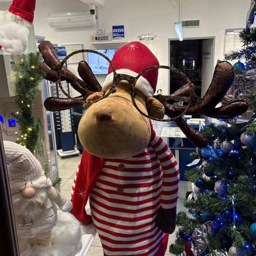 Ein weihnachtlicher Plüsch-Rentier mit Brille und rotem und weißem gestreiftem Pullover steht neben einem Weihnachtsbaum mit blauen Kugeln. Dahinter befindet sich ein Weihnachtsmann und ein Schild mit der Aufschrift 'ZEIS'.