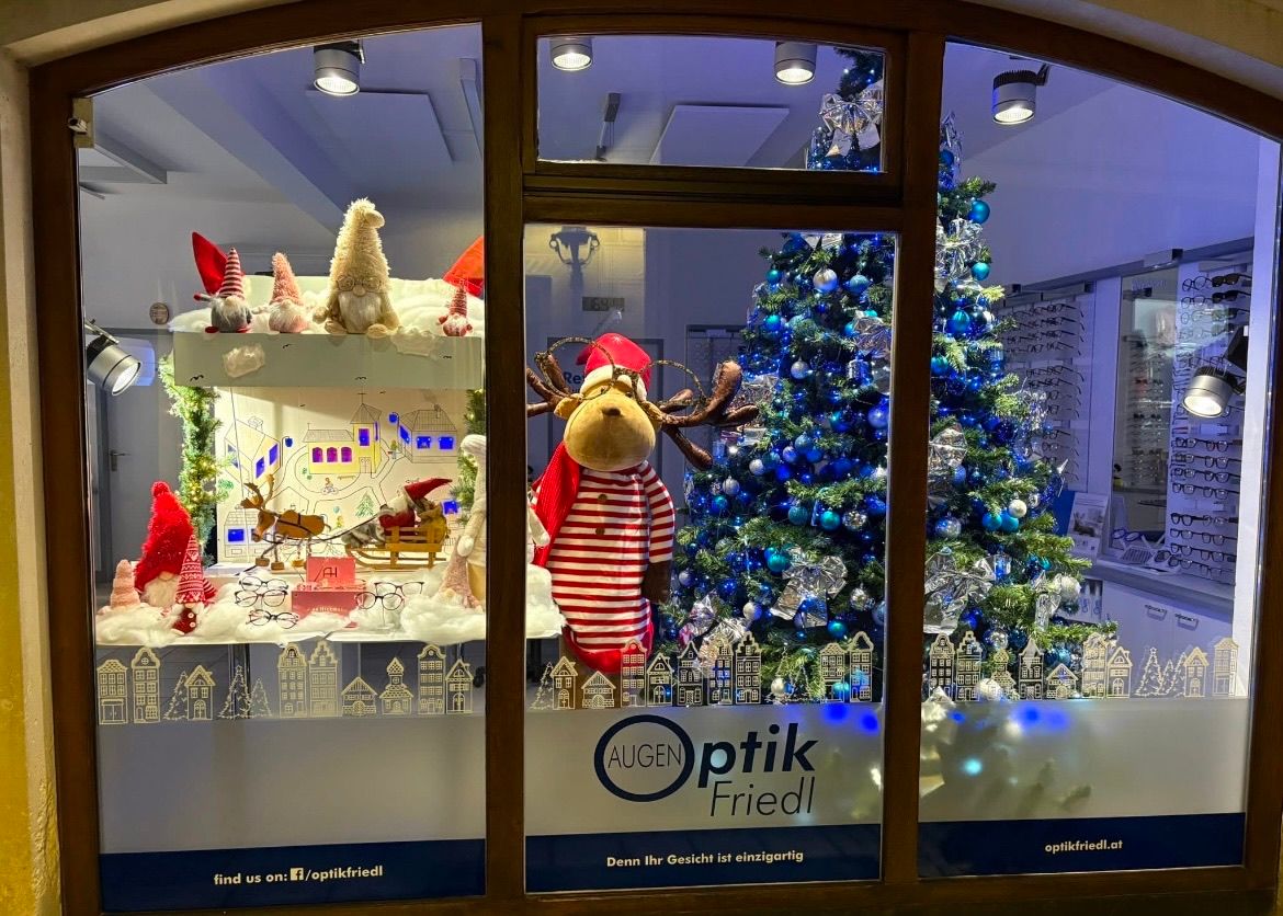 Ein Schaufenster mit einem Rentier in einem Weihnachtsmannhut und gestreiftem Hemd. Das Fenster zeigt auch Weihnachtsdekorationen, darunter Zwerge und einen Schlitten, und einen Weihnachtsbaum mit blauen Ornamenten. Ein Schild sagt 'AUGEN Optik Friedl'.
