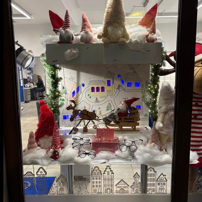 Ein Fensterdisplay zeigt eine verschneite Landschaft mit Zwergen und einem Schlitten, Weihnachtsdekorationen und Brillen auf einem Tisch. Die Zwerge tragen Mützen, und der Schlitten ist mit Rentieren und dem Weihnachtsmann verziert. Hinter dem Display sitzt eine Frau am Schreibtisch.