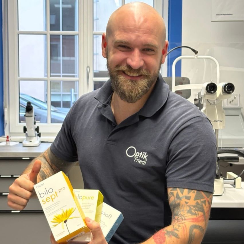 A man with a tattoo smiles and gives a thumbs up while holding two eye care product boxes. He stands in an eye care clinic.