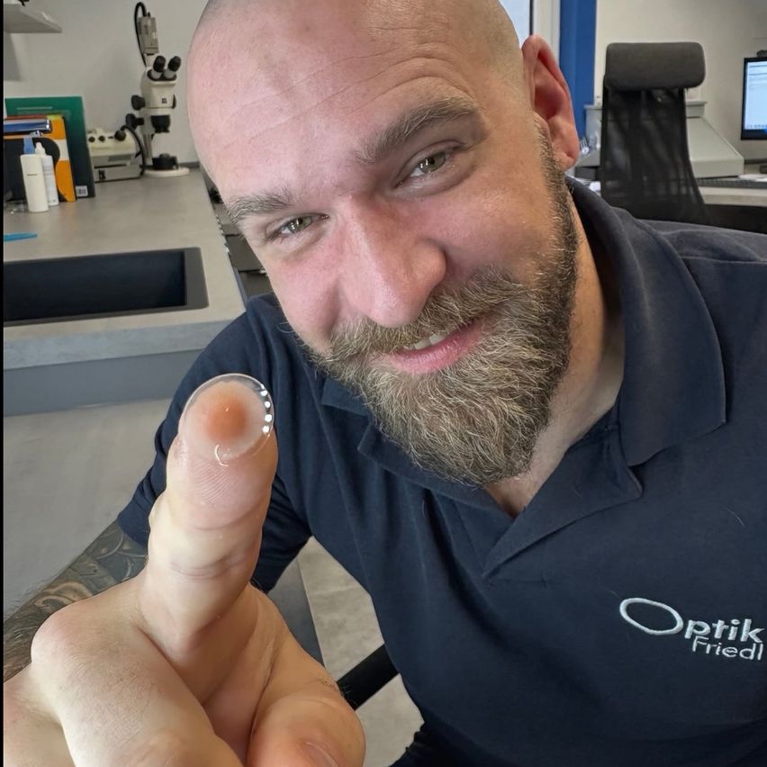 A bearded man wearing a blue polo shirt with a logo holds a contact lens on his finger in an office.
