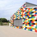 Ein farbenfrohes Gebäude mit Lego-ähnlicher Fassade steht in einem offenen Bereich mit Bäumen und einem Zaun. Ein Rollladen bedeckt ein Fenster.