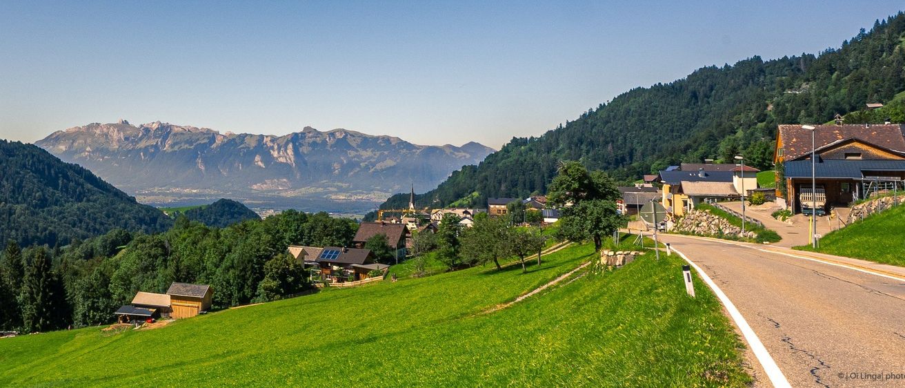 Ein Dorf in den Bergen, umgeben von üppigem Grün. Einige Häuser mit Solarpanelen, ein Turm und eine Bergkette im Hintergrund.