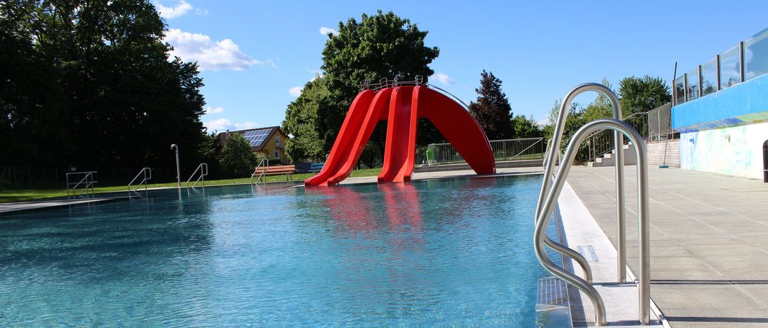 Ein großer Swimmingpool mit einem roten Wasserrutsche an der Seite. Der Pool ist von Bäumen umgeben und hat klares blaues Wasser.