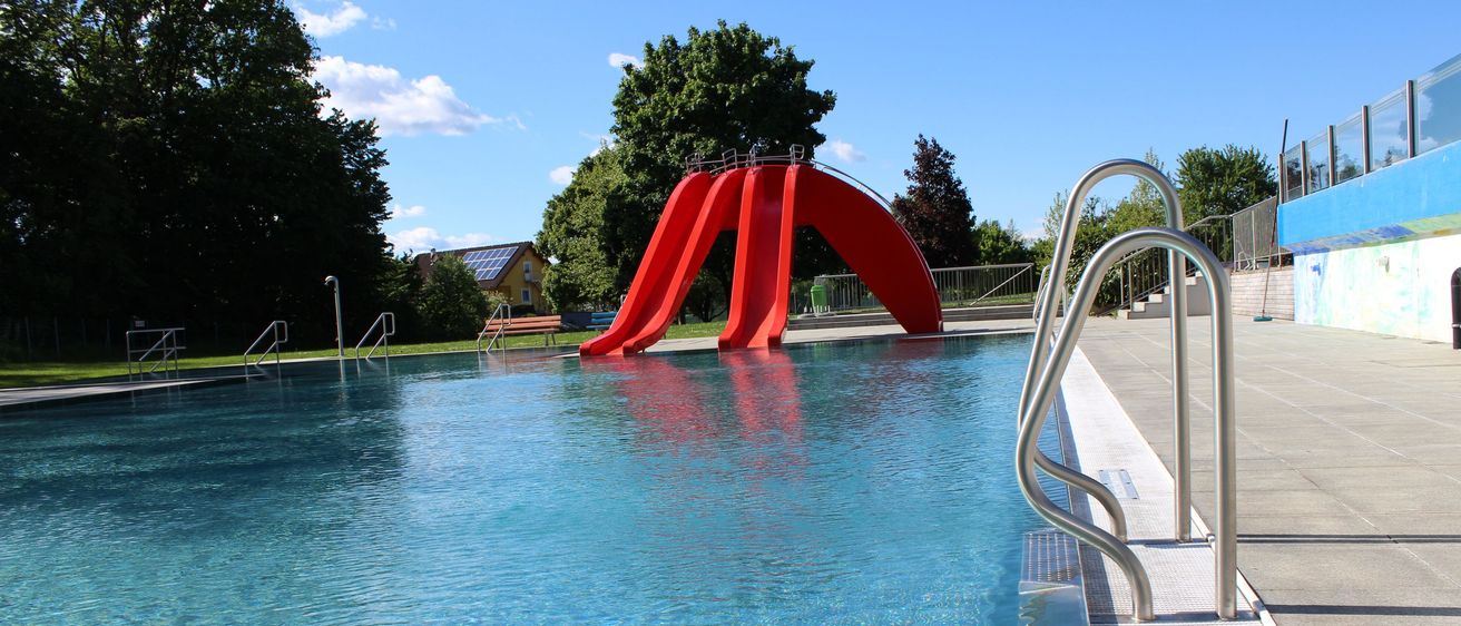 Ein großer Swimmingpool mit einem roten Wasserrutsche an der Seite. Der Pool ist von Bäumen umgeben und hat klares blaues Wasser.