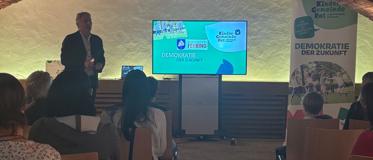 Ein Mann steht vor einem Bildschirm mit einer Präsentation. Dahinter ein Ziegelbogen. Ein Publikum sitzt vor ihm. Ein Banner steht Demokratie der Zukunft.