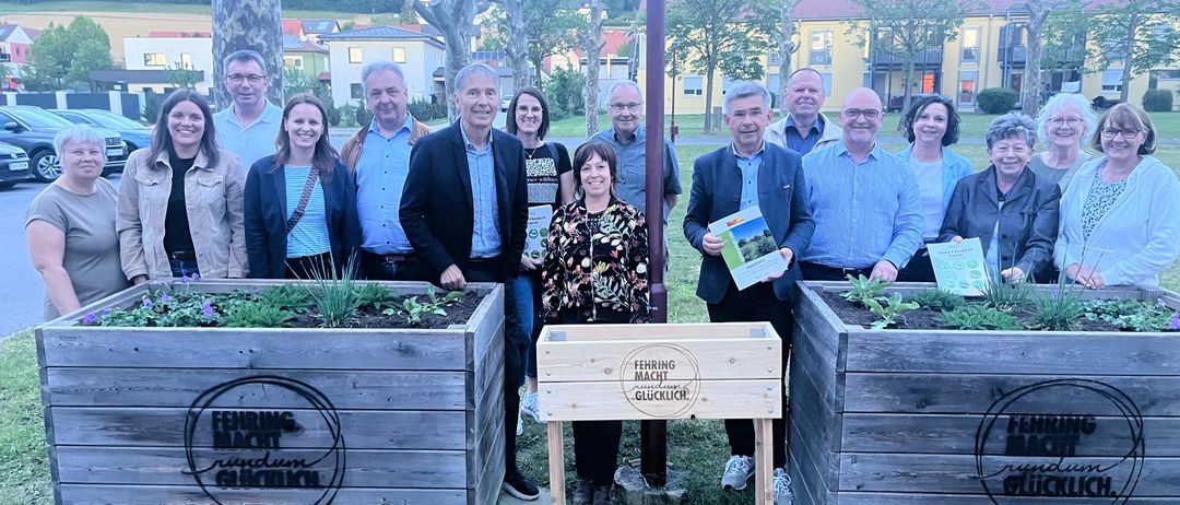 Eine Gruppe von Menschen steht um Holzpflanzer mit Pflanzen herum, lächelt und hält ein Buch. Im Hintergrund sind Gebäude und Bäume sichtbar.