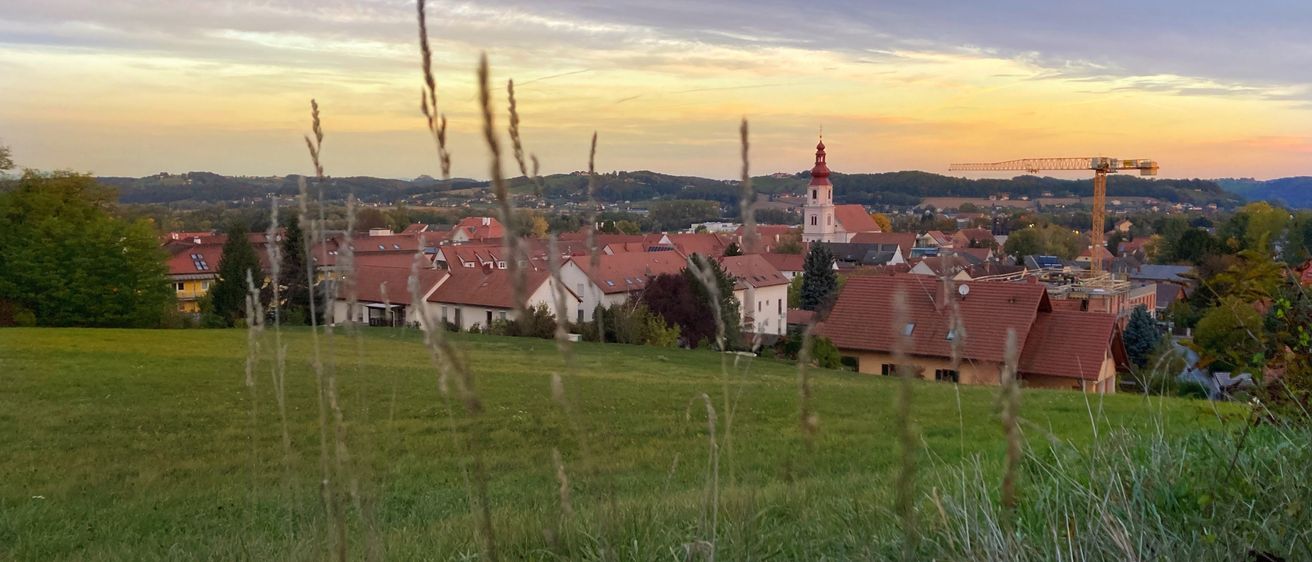Eine Stadt mit roten Dächern und einem Turm unter einem farbenfrohen Sonnenuntergangshimmel. Die Stadt ist von grünen Feldern und Bäumen umgeben.