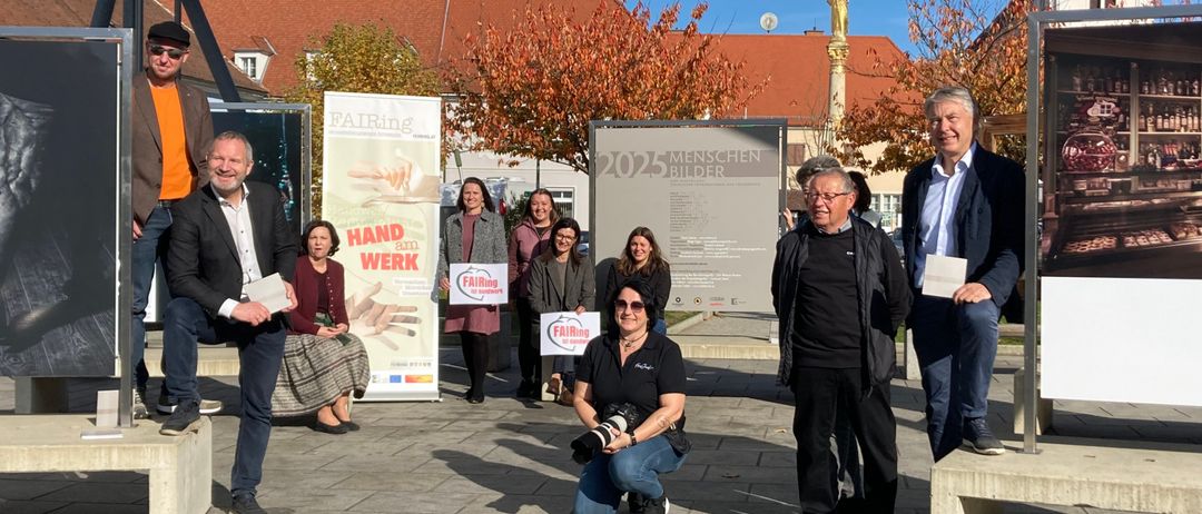 Eine Gruppe von Menschen steht auf einem Platz. Eine Person macht ein Foto. Sie halten alle Schilder und tragen Winterkleidung. Hinter ihnen steht ein Poster mit den Worten 2025 Menschen Bilder.