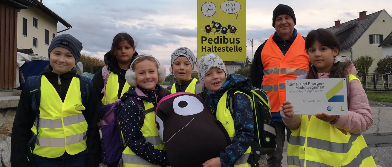 Eine Gruppe von Kindern und einem Mann in Warnwesten stehen draußen. Die Kinder halten ein Schild für Klima-und Energie-Modellregionen.