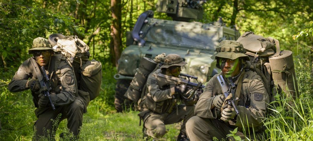 Soldaten in Militäruniformen kauen im Wald, einer hält eine Waffe, der andere ein Gewehr. Ein Militärfahrzeug mit einer angebrachten Waffe steht hinter ihnen.