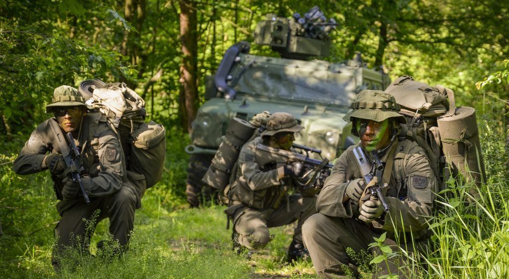 Soldaten in Militäruniformen kauen im Wald, einer hält eine Waffe, der andere ein Gewehr. Ein Militärfahrzeug mit einer angebrachten Waffe steht hinter ihnen.