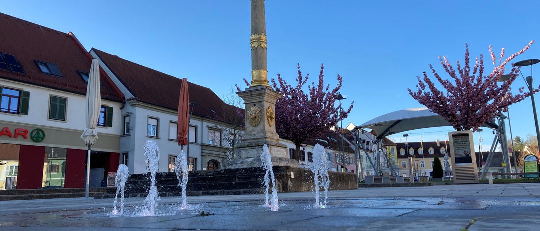 Eine goldene Statue auf einer hohen Säule steht auf einem Platz mit Wasserbrunnen, umgeben von Gebäuden und Bäumen unter einem klaren blauen Himmel.