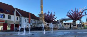 Eine goldene Statue auf einer hohen Säule steht auf einem Platz mit Wasserbrunnen, umgeben von Gebäuden und Bäumen unter einem klaren blauen Himmel.