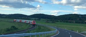Ein roter Zug fährt auf den Gleisen in einem ländlichen Gebiet. Die Straße ist leer mit einem Geländer auf der Seite. Berge und grüne Felder sind in der Ferne sichtbar.
