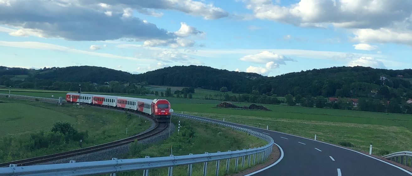Ein roter Zug fährt auf den Gleisen in einem ländlichen Gebiet. Die Straße ist leer mit einem Geländer auf der Seite. Berge und grüne Felder sind in der Ferne sichtbar.