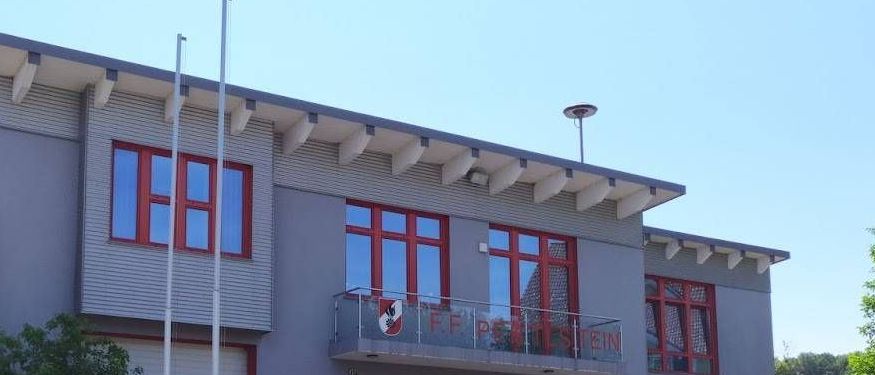 Ein modernes Feuerwehrhaus mit roten Akzenten und Glasfenstern, mit einem Balkon, einem roten Schild und einem Fahnenmast davor.