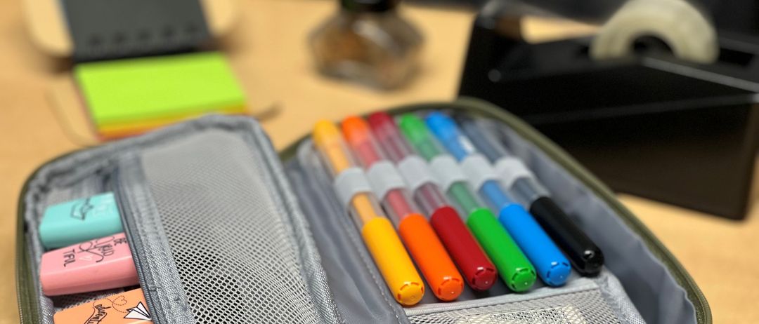 Eine offene Stiftetasche mit mehreren farbigen Stiften und Markern darin, auf einem Holztisch.