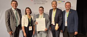 Vier Personen stehen bei einer Preisverleihung zusammen, einer Mann hält ein Zertifikat und ein anderer einen Pokal. Sie tragen alle Namensschilder.