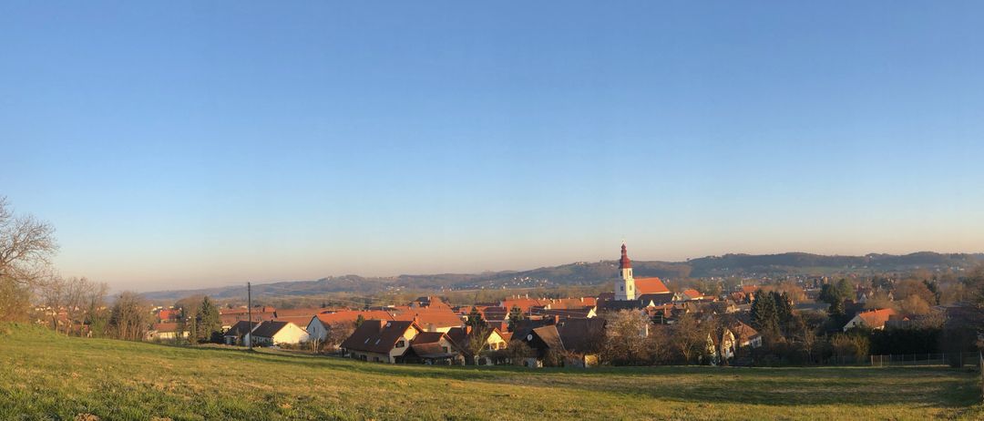 Ein Panoramablick auf ein Dorf mit rot gedeckten Häusern unter einem klaren blauen Himmel. Das Dorf ist von Grün umgeben und in der Ferne befinden sich Berge.