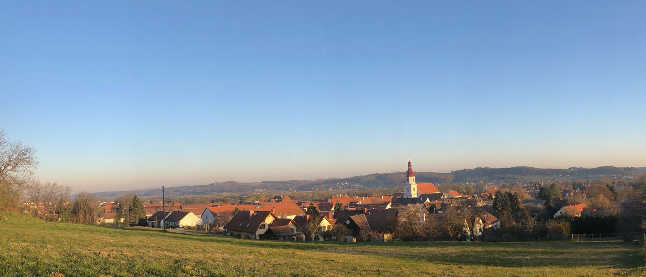Ein Panoramablick auf ein Dorf mit rot gedeckten Häusern unter einem klaren blauen Himmel. Das Dorf ist von Grün umgeben und in der Ferne befinden sich Berge.