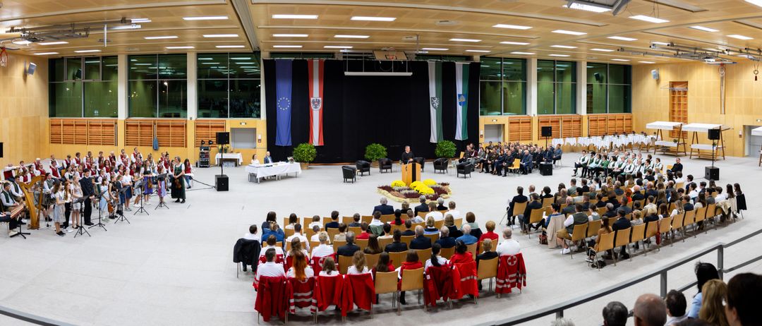 Eine offizielle Veranstaltung in einem großen Saal mit Fahnen und Leuten, die auf Stühlen sitzen. Ein Podium steht in der Mitte, an dem eine Person spricht. Einige Personen tragen rote Jacken.
