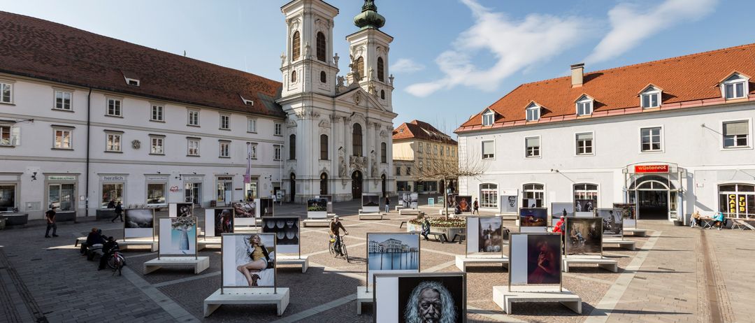 Ein Platz mit Kopfsteinpflaster zeigt zahlreiche Fotografien vor einem historischen Gebäude unter einem klaren Himmel.