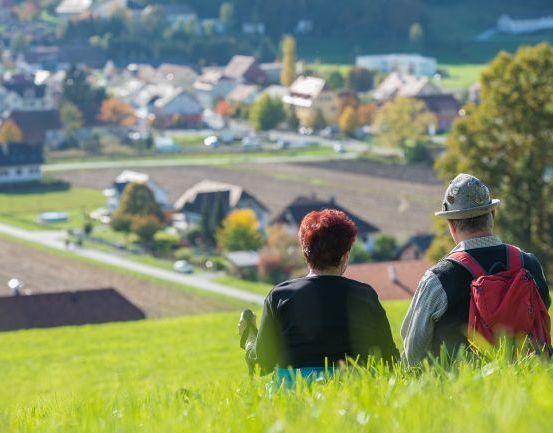 Ein älteres Paar sitzt auf einem grünen Hügel und blickt auf ein Dorf mit mehreren Häusern und Bäumen.