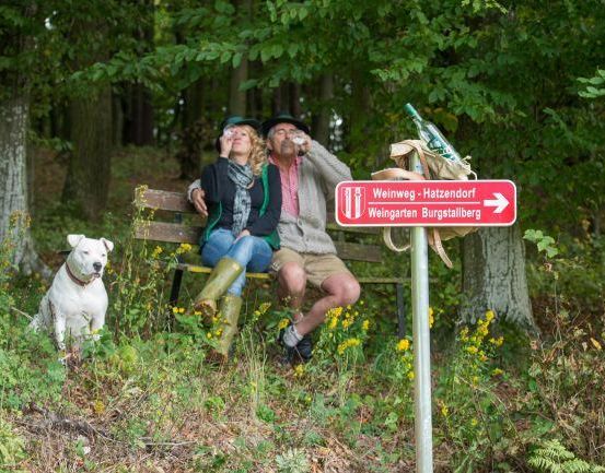 Zwei Personen sitzen auf einer Bank im Wald und trinken aus Flaschen. Ein Hund sitzt in der Nähe. Ein Schild weist auf Hatzendorf hin, mit Burgstallberg im Hintergrund.