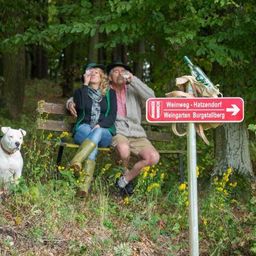 Zwei Personen sitzen auf einer Bank im Wald und trinken aus Flaschen. Ein Hund sitzt in der Nähe. Ein Schild weist auf Hatzendorf hin, mit Burgstallberg im Hintergrund.