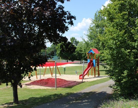 Ein Spielplatz mit Rutsche und Klettergerüst, umgeben von Bäumen und Gras, mit einem Weg, der dorthin führt.