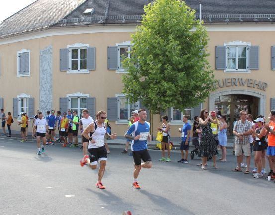 Läufer nehmen an einem Marathon teil, während Zuschauer von der Seite anfeuern, vor einem Gebäude mit dem Schild 'EUERWEHR'.