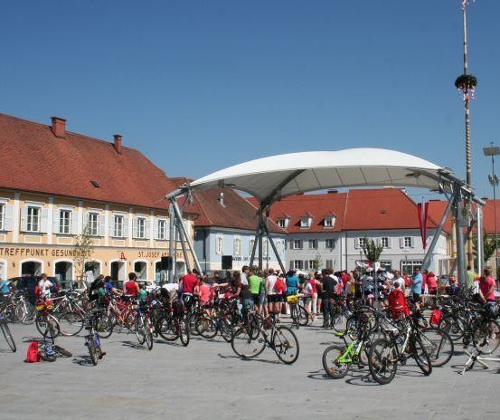 Eine Menschenmenge mit Fahrrädern versammelt sich auf einem Stadtplatz, mit einem Vordach, Gebäuden und Straßenlaternen im Hintergrund.