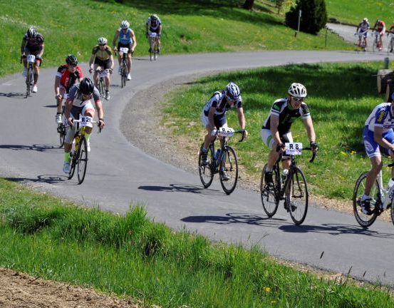 Eine Gruppe von Radfahrern fährt auf einer Straße mit einer Kurve. Sie tragen Helme und Radsportbekleidung. Einige haben Flaschen dabei. Die Straße hat auf beiden Seiten eine Rasenfläche.