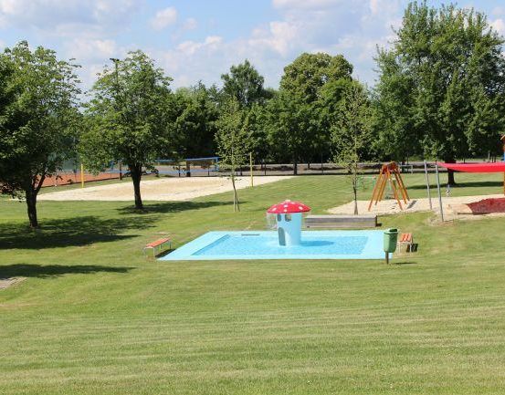 Ein Kinderspielplatz mit einem Brunnen in der Mitte, umgeben von einer Rasenfläche und Bäumen, mit einem Volleyballplatz im Hintergrund.