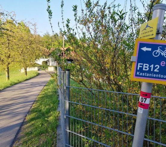 Ein FB12 Kastanientour-Schild ist neben einem Metallzaun montiert, mit Bäumen und einem Haus im Hintergrund.
