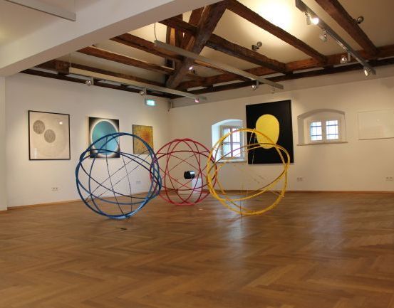 Ein leerer Kunstraum mit Holzböden und weißen Wänden. Mehrere abstrakte Kunstwerke schmücken die Wände, darunter ein gelbes Sonnenstück. Vier farbenfrohe kugelförmige Skulpturen sind auf dem Boden platziert.